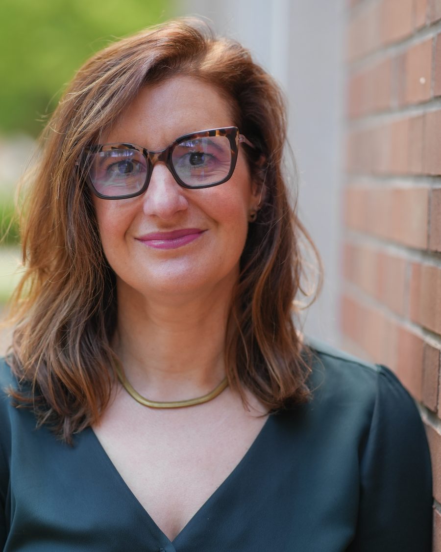 Claire Kamp Dush, smiling, in tortoiseshell glasses beside a brick wall